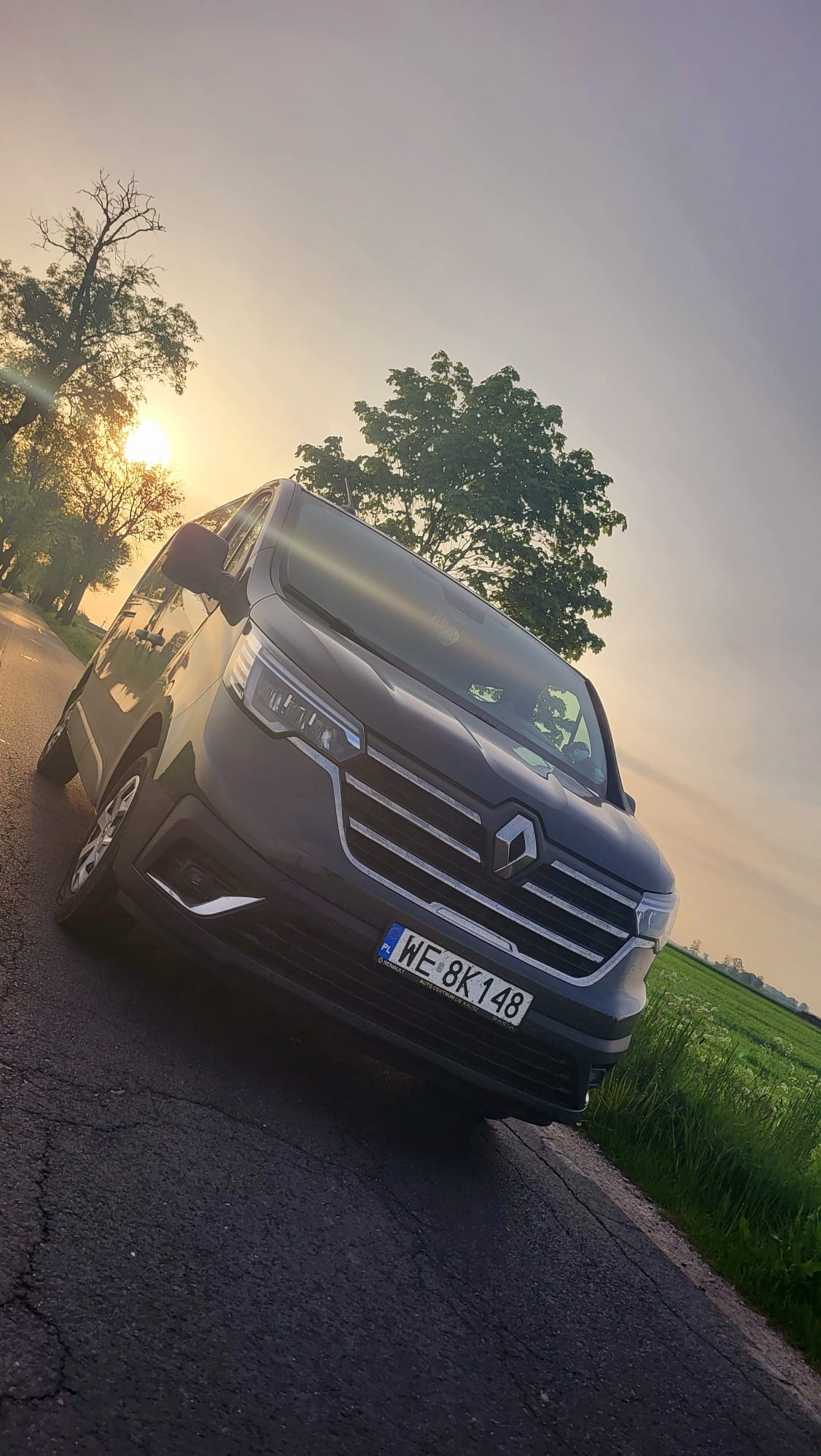 Szary Renault Trafic z polską tablicą rejestracyjną WE 8K148, zaparkowany na poboczu drogi polnej o zachodzie słońca.