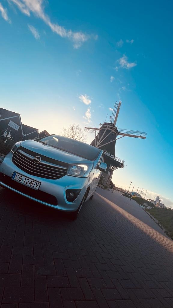 Srebrny Opel Vivaro z bydgoską rejestracją CB-124EV na tle wiatraka i błękitnego nieba.