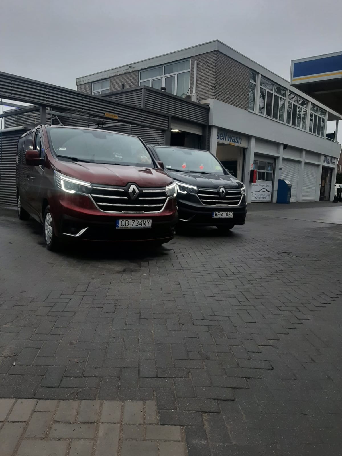 Dwa busy Renault, bordowy i czarny, zaparkowane na brukowanym placu przed myjnią samoobsługową w pochmurny dzień.