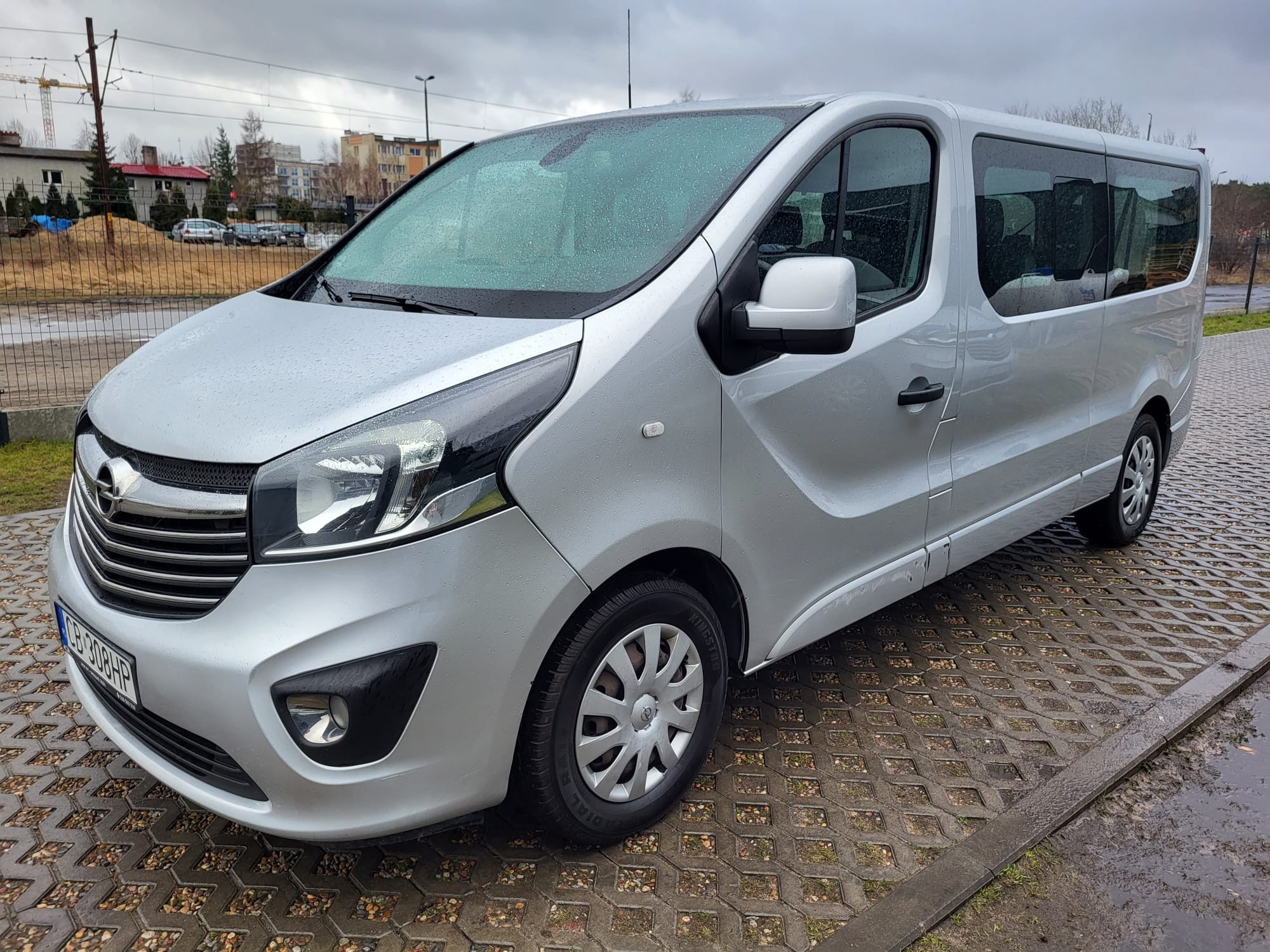 Srebrny Opel Vivaro z widoczną tablicą rejestracyjną, zaparkowany na betonowej kostce w pochmurny dzień.
