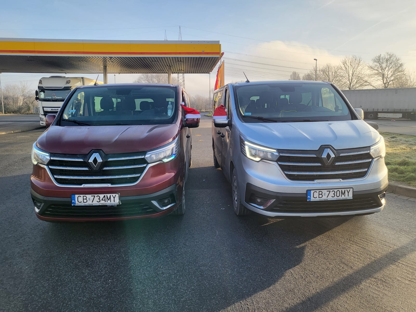 Dwa busy Renault, bordowy i srebrny, zaparkowane obok siebie na stacji benzynowej, oba z czapkami Mikołaja na lusterkach.