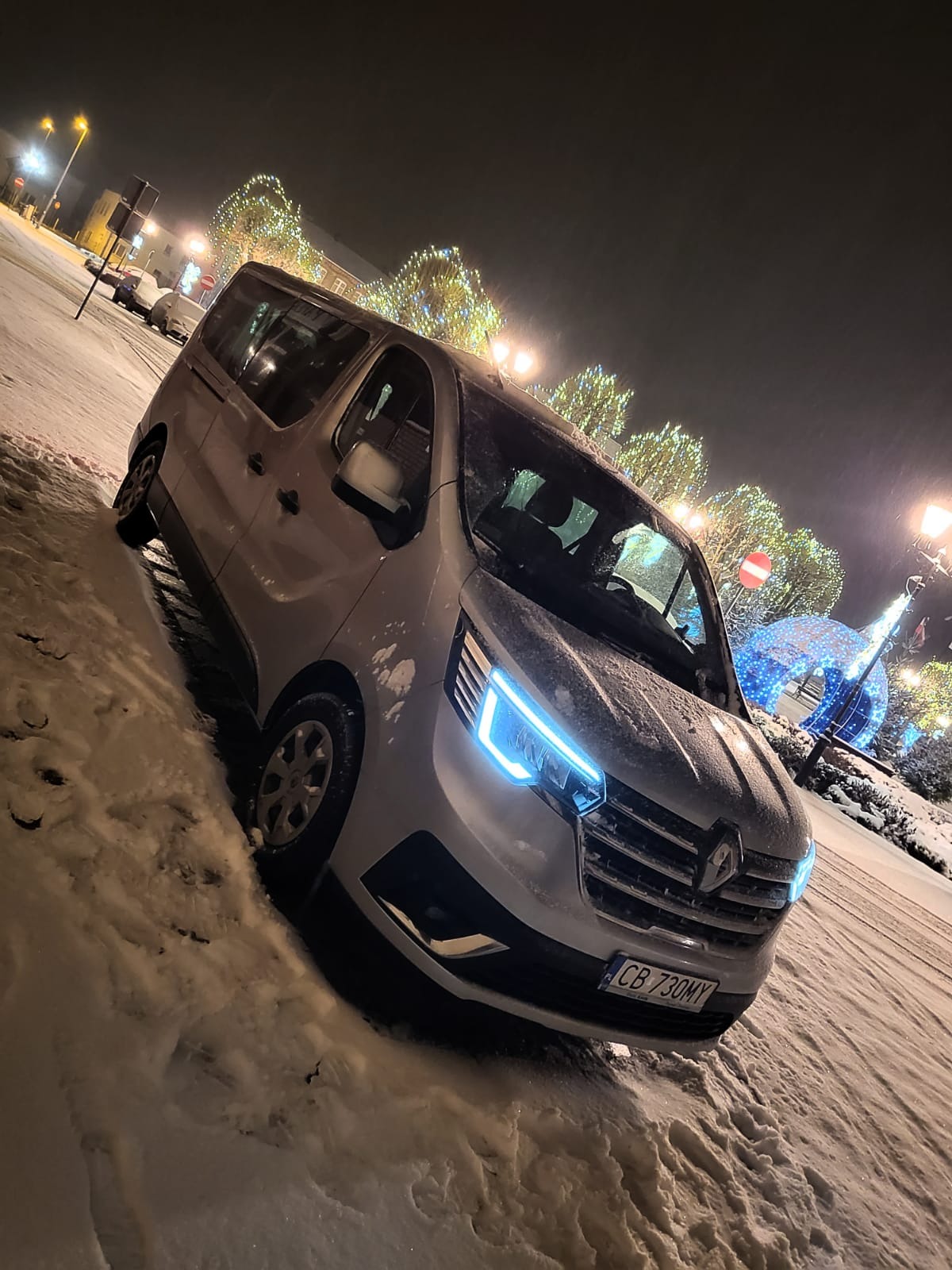 Srebrny van Renault zaparkowany w śniegu nocą, z włączonymi niebieskimi światłami LED, na tle oświetlonego miejskiego placu.