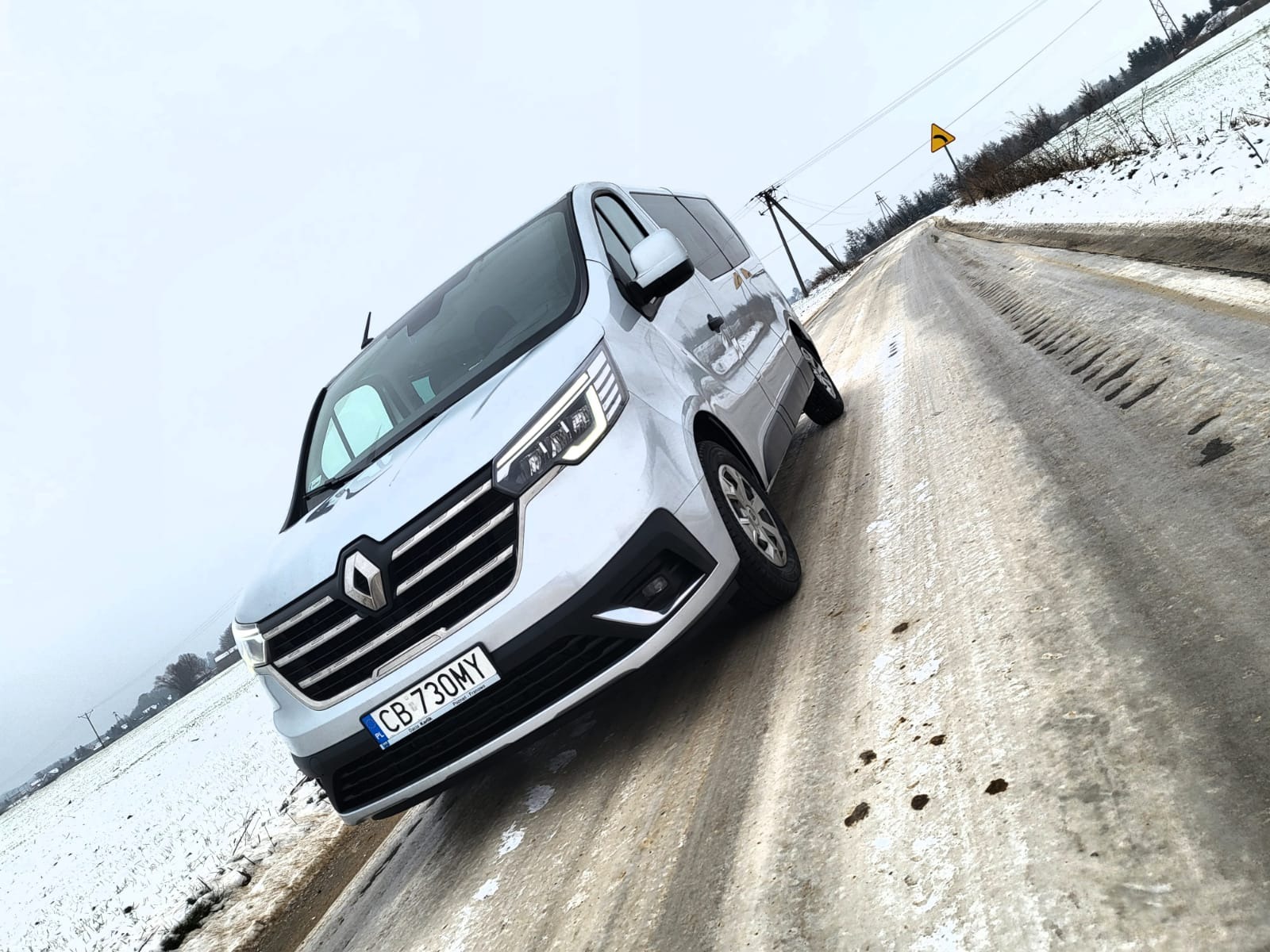 Srebrny Renault Trafic z polską tablicą rejestracyjną CB 730MY na ośnieżonej drodze wiejskiej w pochmurny dzień.