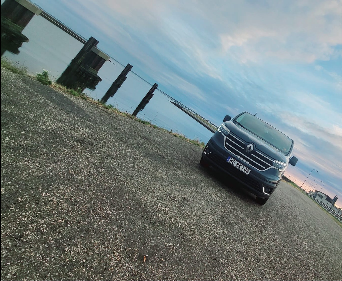 Ciemnoniebieski Renault Trafic z polską tablicą rejestracyjną WE 8K148 zaparkowany na asfalcie, w tle widok na jezioro i drewniane pale wbite w wodę, pochmurne niebo.