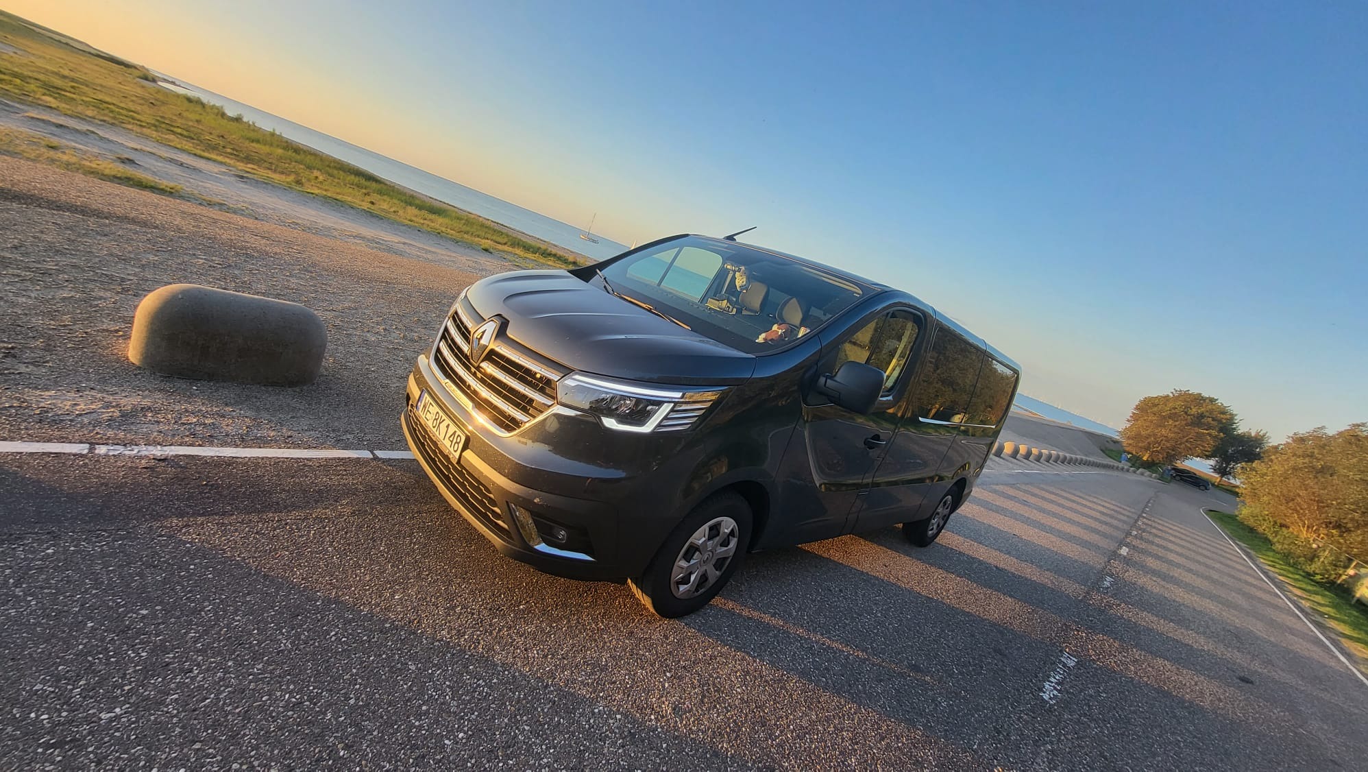 Czarny bus Renault Trafic zaparkowany na asfalcie z widokiem na morze i zachodzące słońce, zarys żaglówki na horyzoncie.