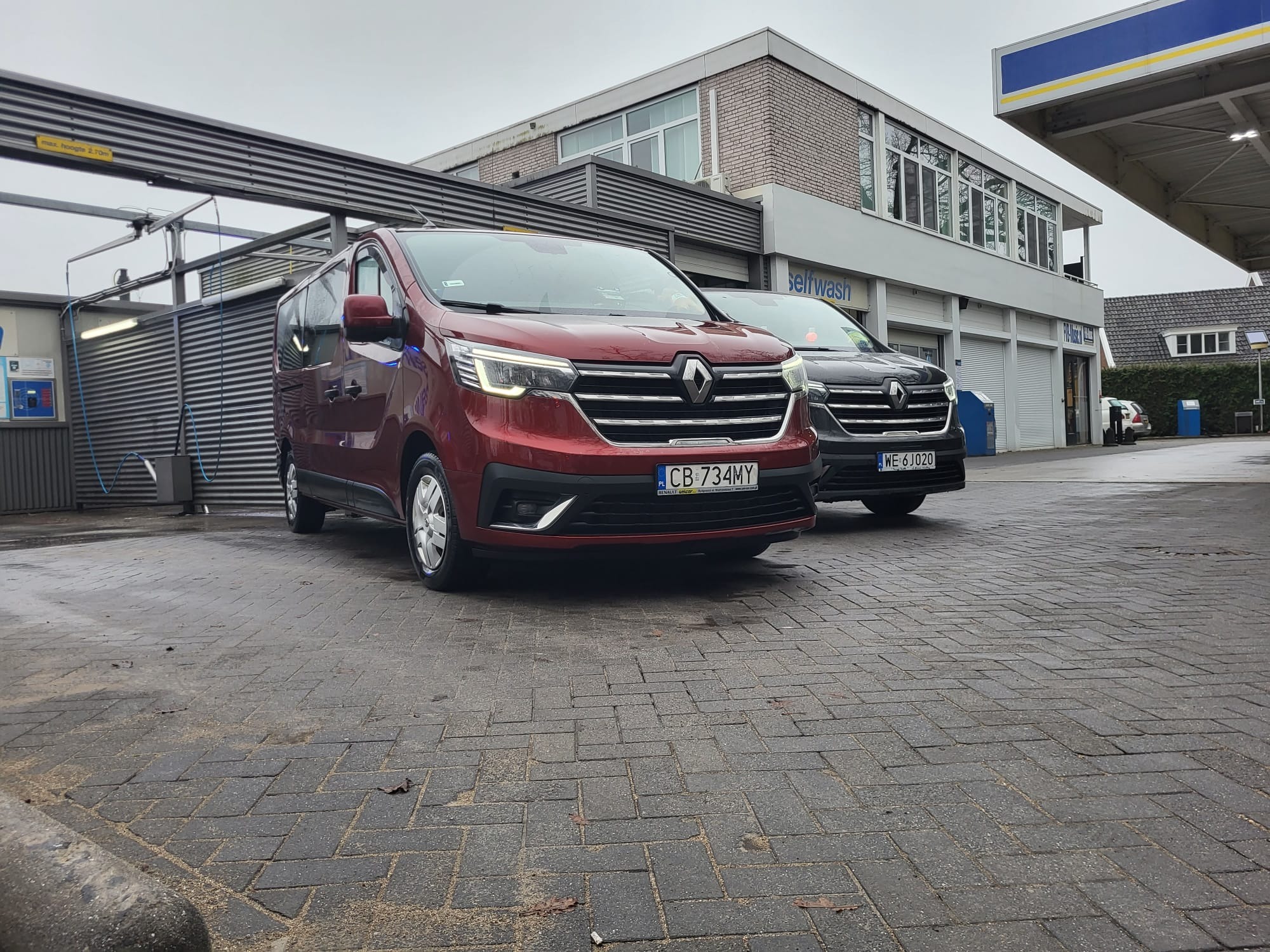 Dwa busy Renault Trafic zaparkowane przed myjnią samochodową, jeden bordowy z polską rejestracją, drugi ciemny z zagraniczną.