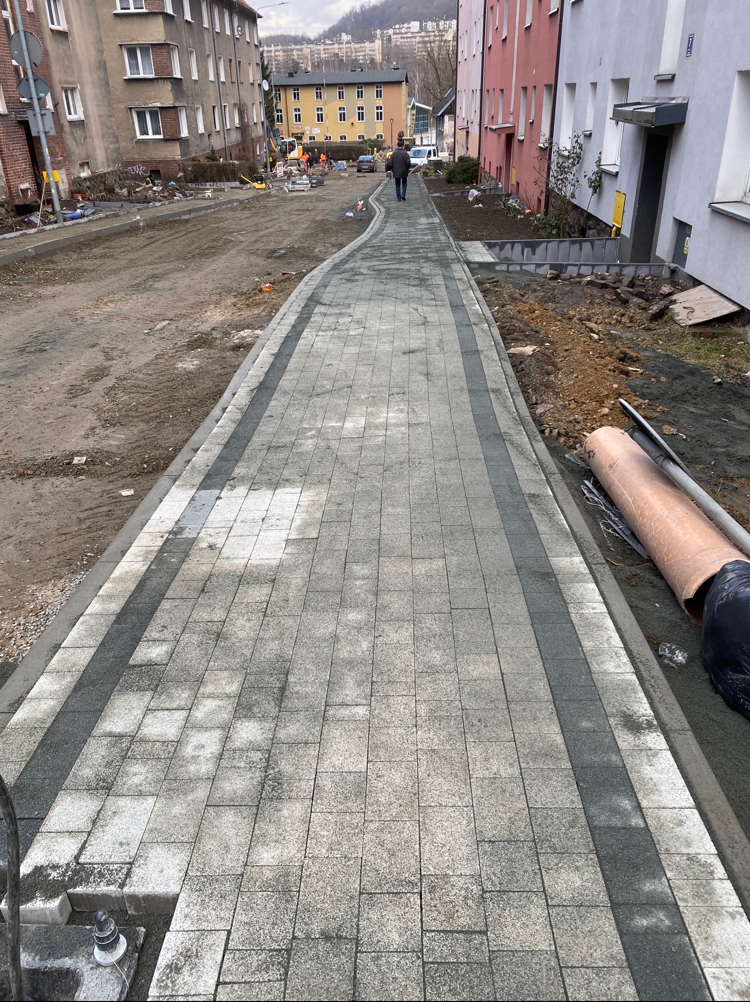 Nowo ułożony chodnik z kostki brukowej w szarych odcieniach z widocznymi pracami ziemnymi w tle oraz robotnikami w oddali.