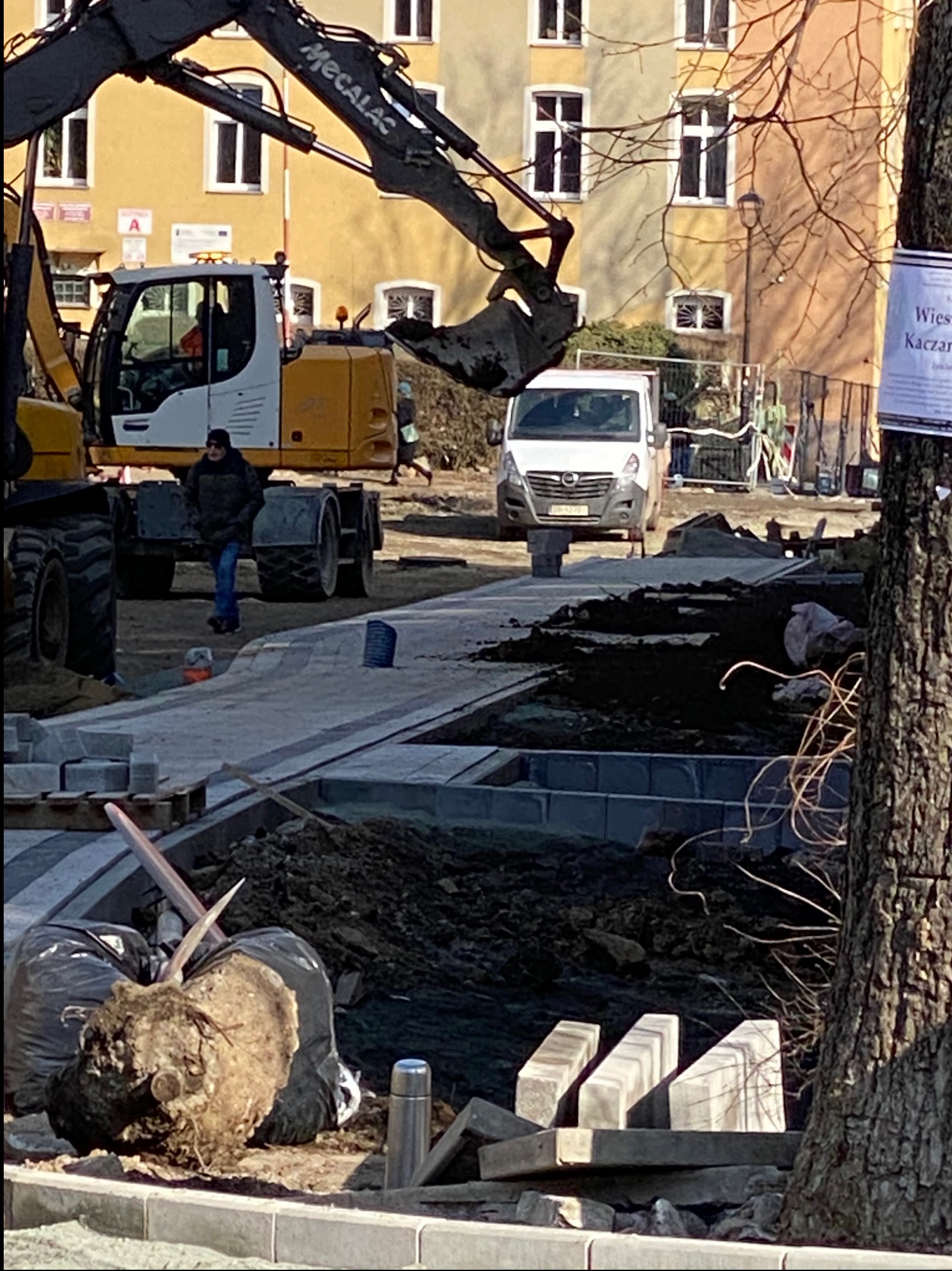 Prace ziemne przy układaniu chodnika z kostki betonowej, w tle koparka Mecalac, biały van i budynek z żółtą elewacją.