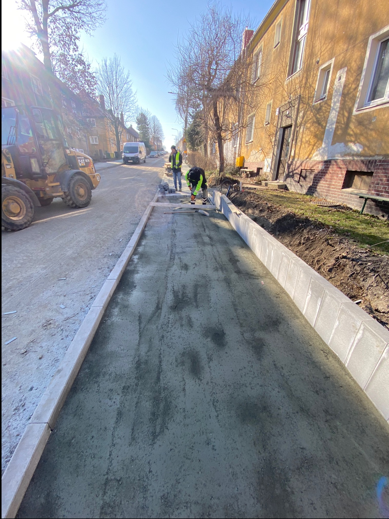 Układanie betonowej nawierzchni chodnika z obrzeżami, widoczny pracownik używający ubijarki oraz koparka CAT w tle.
