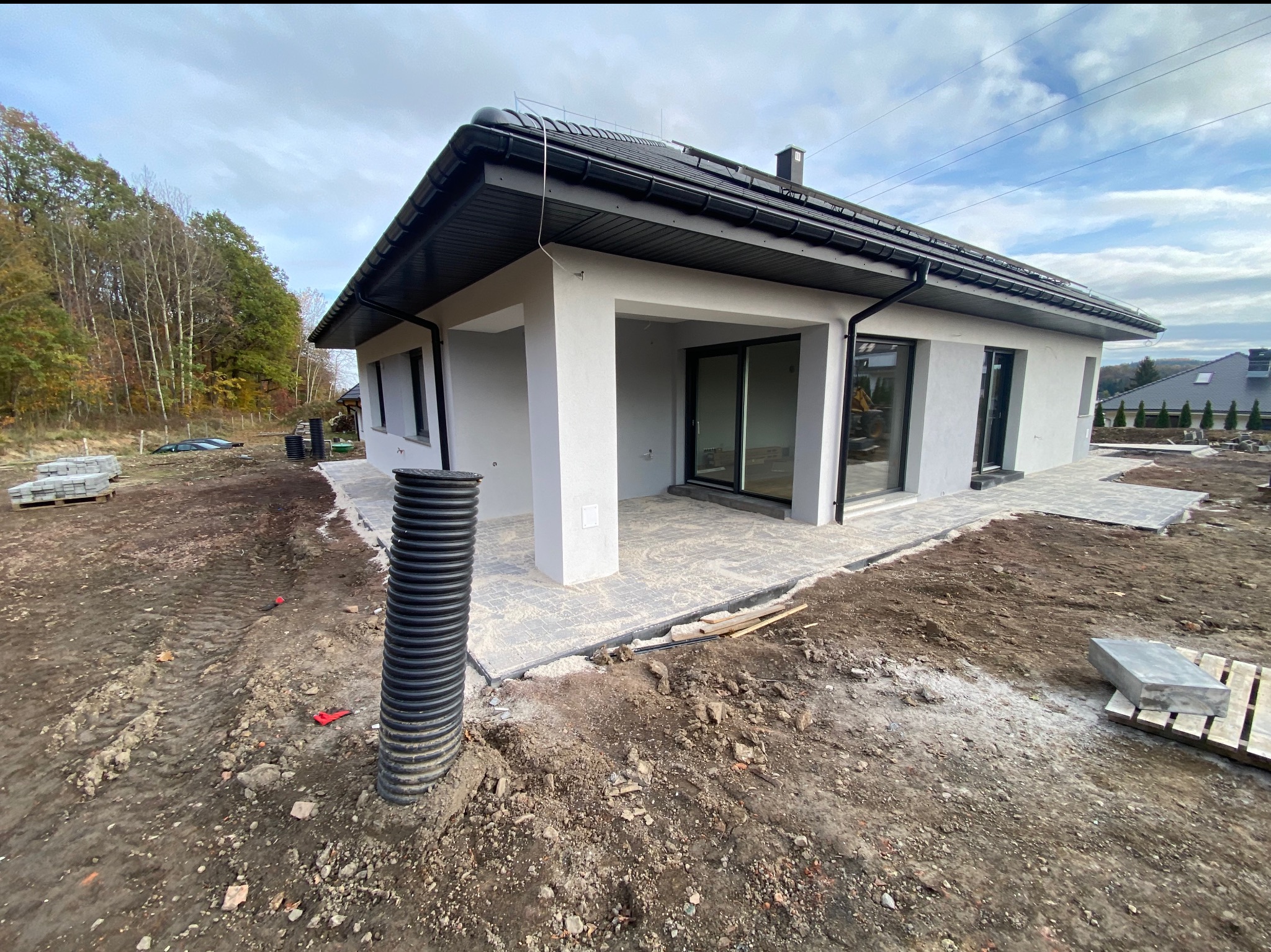 Nowoczesny dom jednorodzinny z szarą elewacją i czarnym dachem, widok z boku na taras z przesuwnymi drzwiami, teren wokół w trakcie zagospodarowania, widoczna rura drenażowa i paleta z kostką brukową.