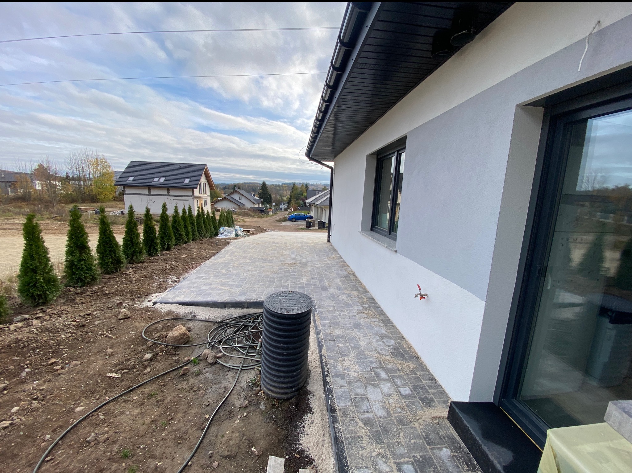 Nowoczesny dom z wybrukowanym podjazdem i rzędem tuj, widoczne przyłącze wody na elewacji.