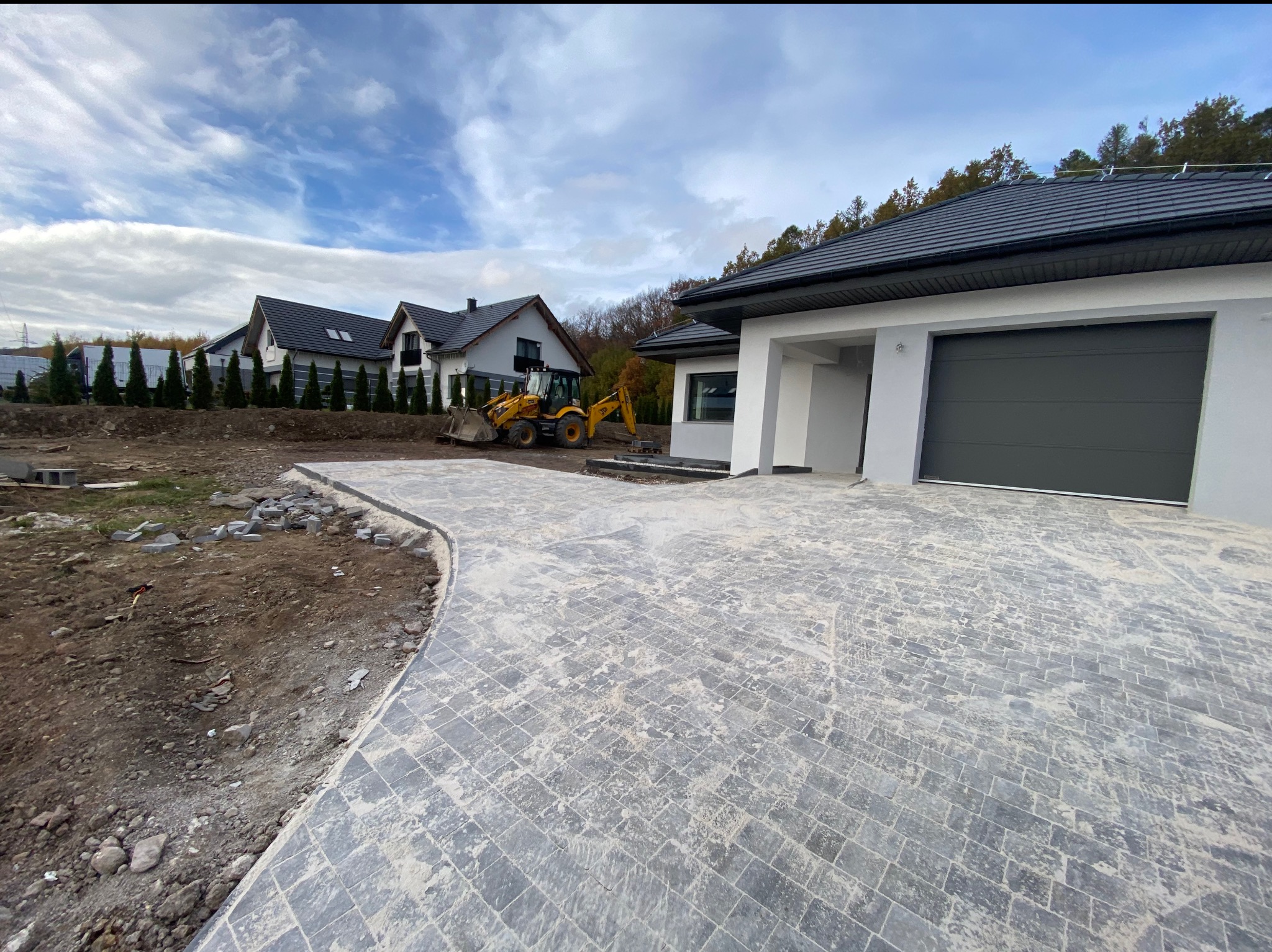 Nowoczesny dom jednorodzinny z szarą bramą garażową i wybrukowanym podjazdem z kostki w romby, w tle koparka i inne domy w trakcie budowy.