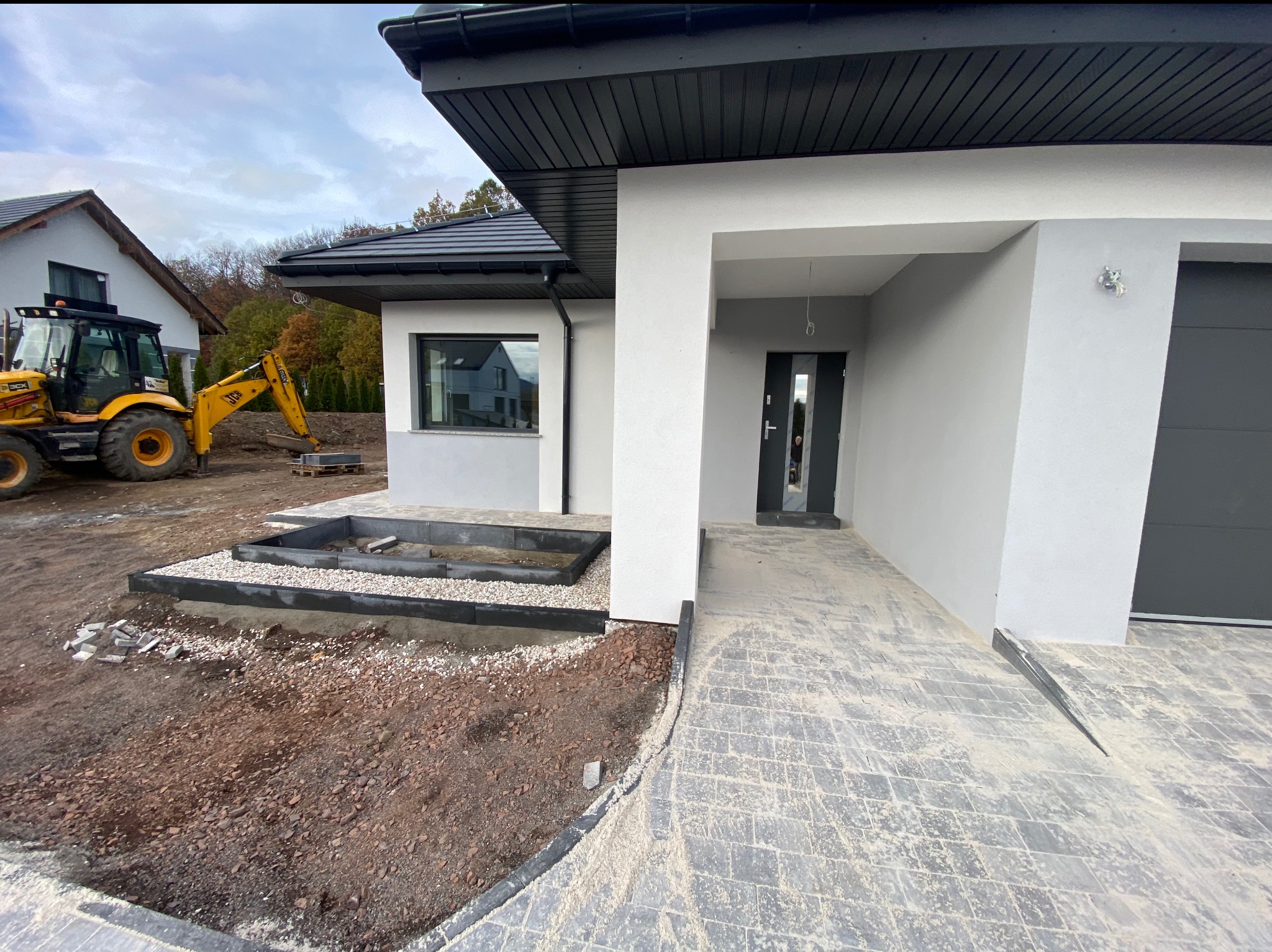 Nowoczesny dom jednorodzinny w trakcie wykańczania ogrodu, widoczny żółty traktor JCB, przygotowane rabaty z kamieniami i obrzeżami, szara kostka brukowa na podjeździe.