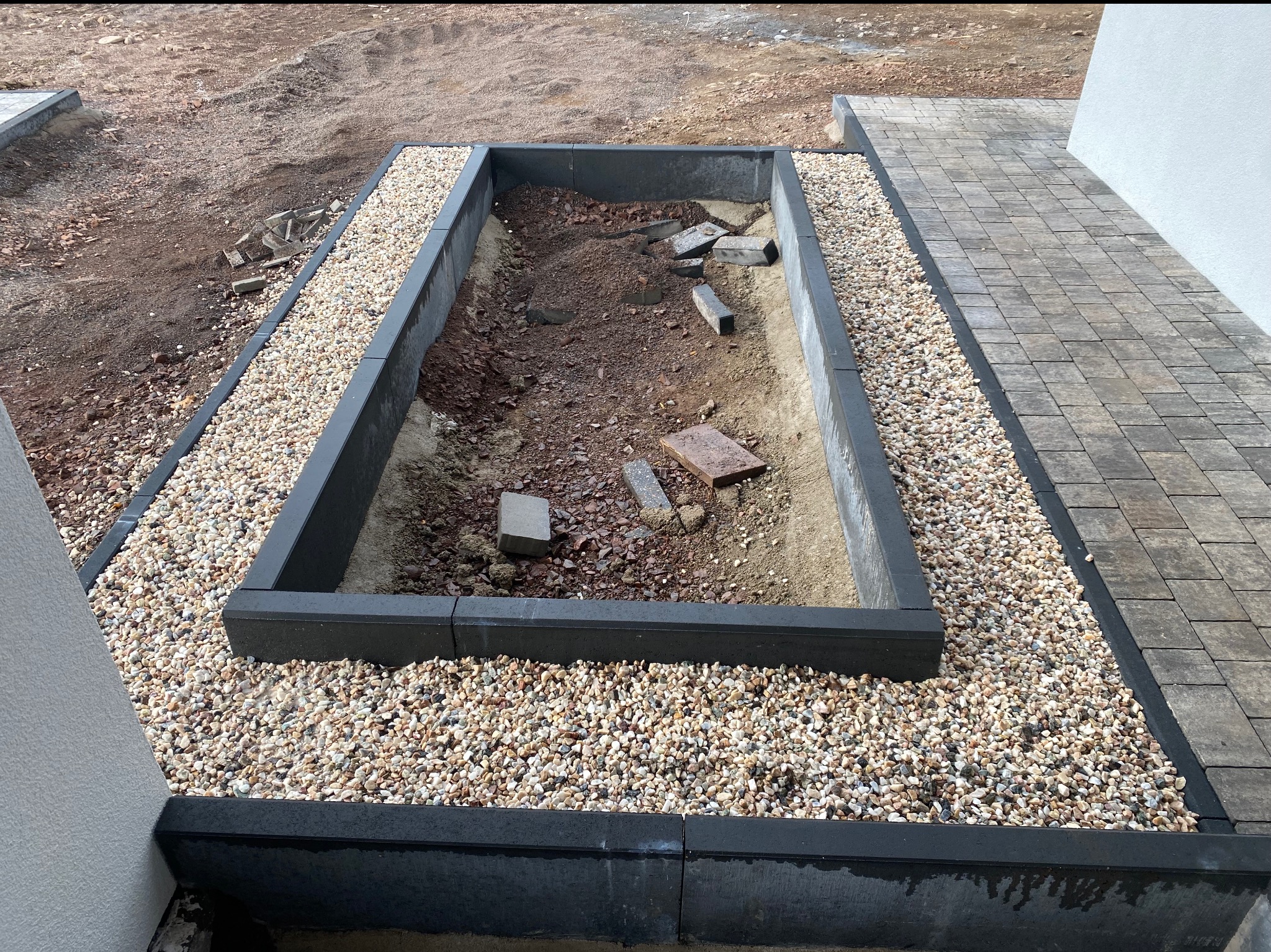 Przygotowanie pod rabatę kwiatową z obrzeżami z kostki brukowej i wysypaną warstwą ozdobnych kamyków, widoczne fragmenty gruzu wewnątrz rabaty.