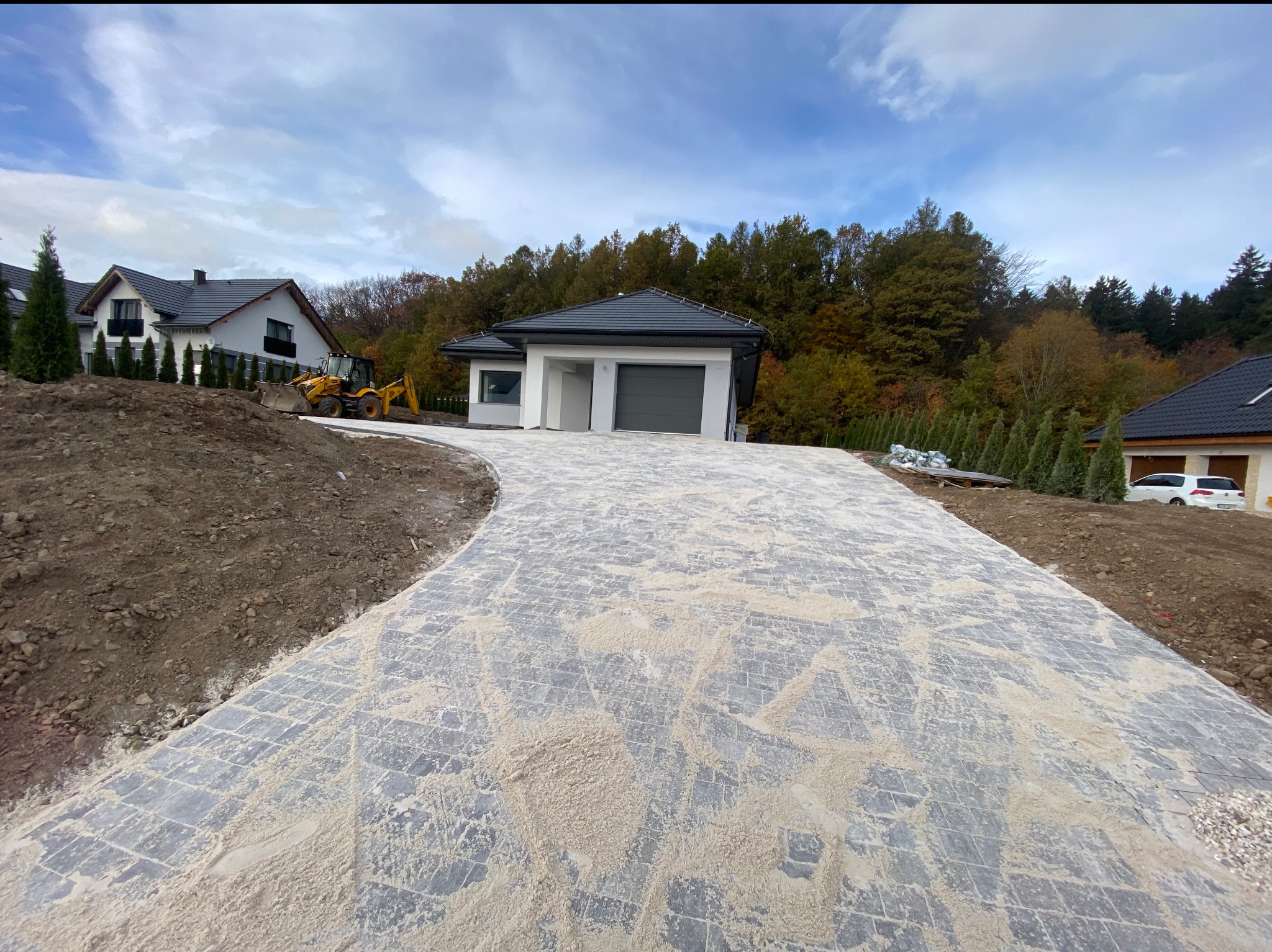 Nowy dom jednorodzinny z szarą kostką brukową na podjeździe, częściowo zasypaną piaskiem, z żółtą koparką w tle i jesiennym krajobrazem.