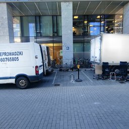 MATEUSZ JEŻEWSKI - Dwa białe busy zaparkowane przed nowoczesnym budynkiem biurowym, obok busa leżą krzesła biurowe, sugerujące przeprowadzkę biura.