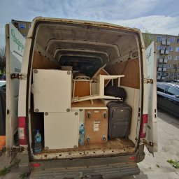 MATEUSZ JEŻEWSKI - Wnętrze białego, częściowo załadowanego busa z otwartymi drzwiami, widoczne meble, walizka i butelki wody mineralnej, w tle blok mieszkalny.