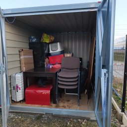 MATEUSZ JEŻEWSKI - Wnętrze otwartego, metalowego garażu typu blaszak wypełnione różnorodnymi przedmiotami: walizka na kółkach, kartony, stół, krzesła w stosie, kuweta dla kota i inne przedmioty zabezpieczone folią.