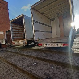 MATEUSZ JEŻEWSKI - Trzy busy do przeprowadzek zaparkowane na schodach z kostki brukowej, jeden z pracownikiem w trakcie rozładunku, widok z dołu pod słońce.