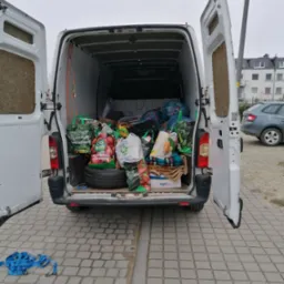 Wnętrze białego busa z otwartymi drzwiami załadowane torbami z zakupami, kocami i oponą, na zewnątrz kostka brukowa i niebieskie pasy transportowe.
