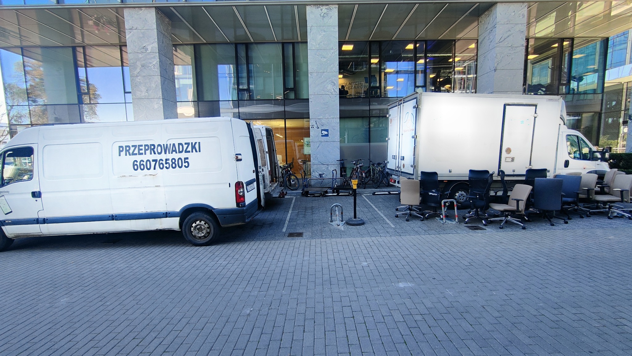 Dwa białe busy zaparkowane przed nowoczesnym budynkiem biurowym, obok busa leżą krzesła biurowe, sugerujące przeprowadzkę biura.