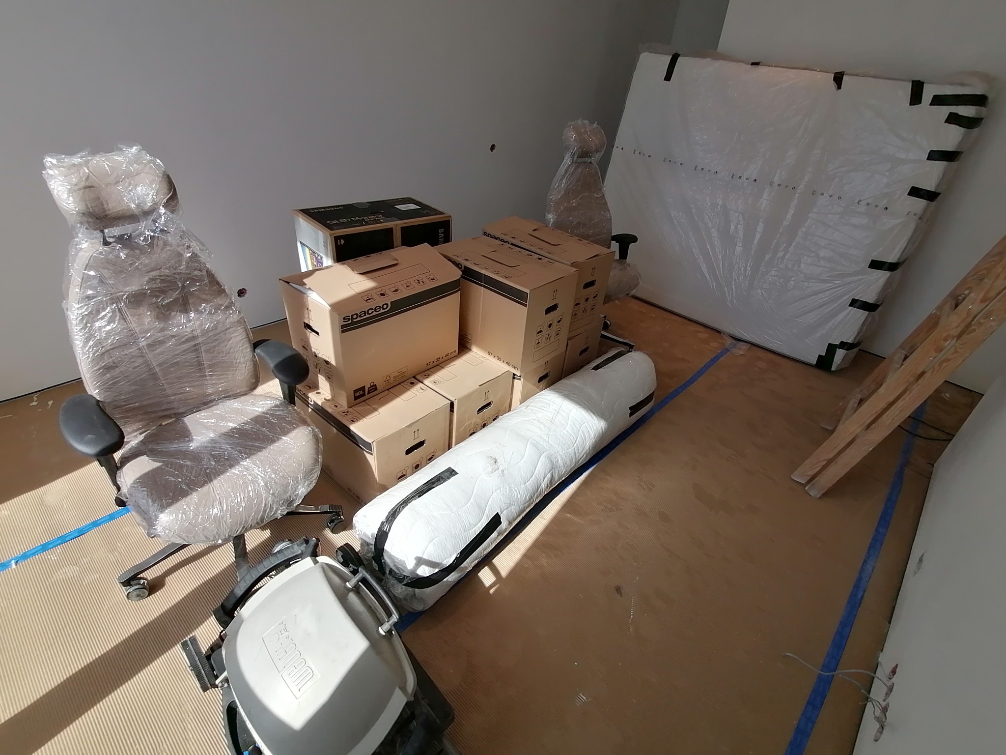 Spakowane meble, krzesło biurowe owinięte folią, kartony Spaceo i materac przygotowane do transportu, widoczne zabezpieczenia podłogi taśmą