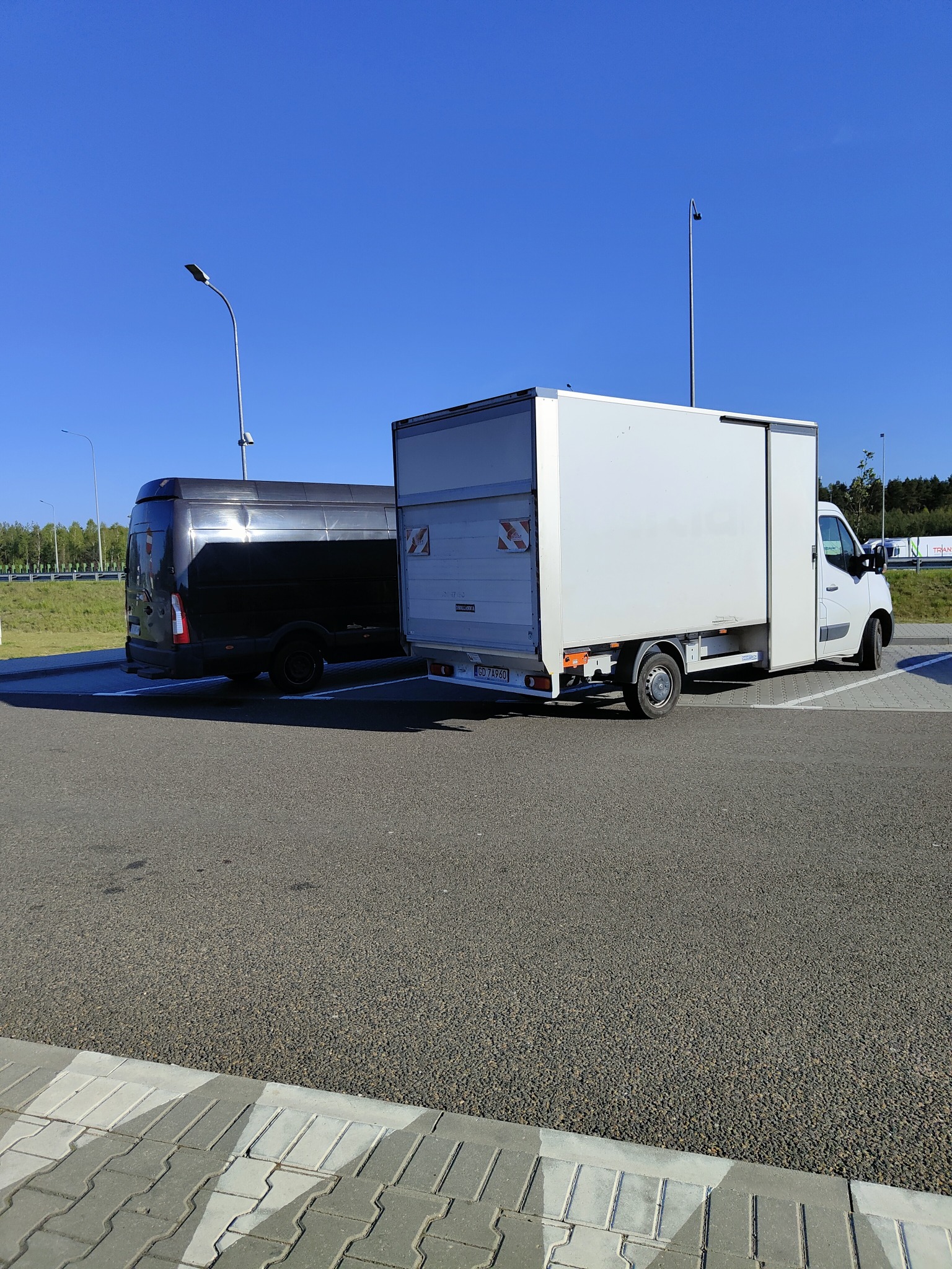 Dwa busy: biały z przesuwanymi drzwiami i czarny, zaparkowane na parkingu z kostki brukowej w słoneczny dzień.