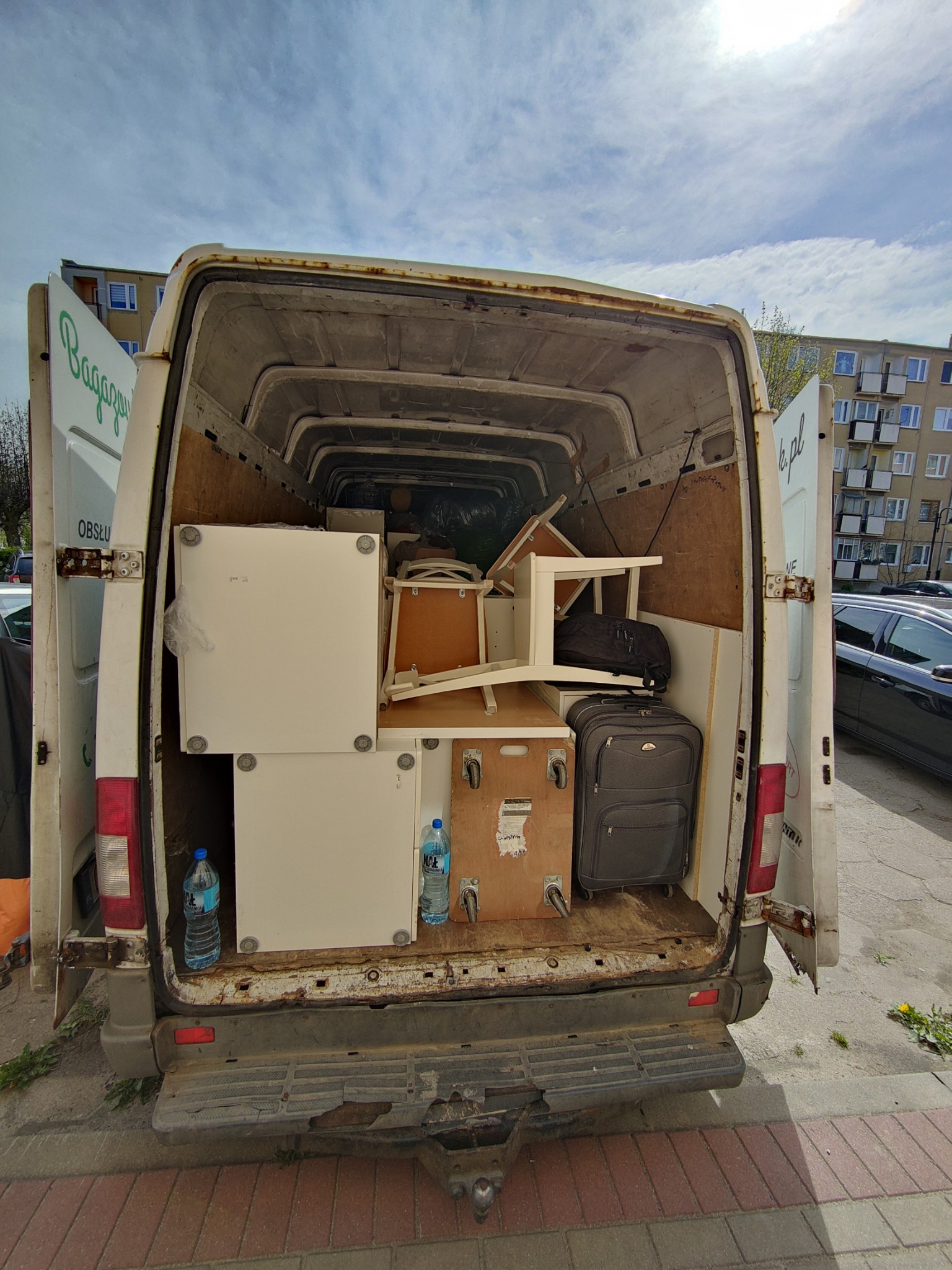 Wnętrze białego, częściowo załadowanego busa z otwartymi drzwiami, widoczne meble, walizka i butelki wody mineralnej, w tle blok mieszkalny.