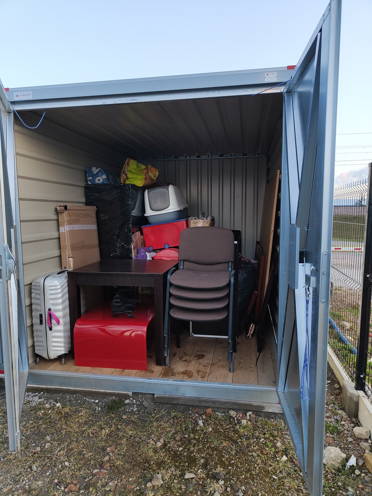 Wnętrze otwartego, metalowego garażu typu blaszak wypełnione różnorodnymi przedmiotami: walizka na kółkach, kartony, stół, krzesła w stosie, kuweta dla kota i inne przedmioty zabezpieczone folią.