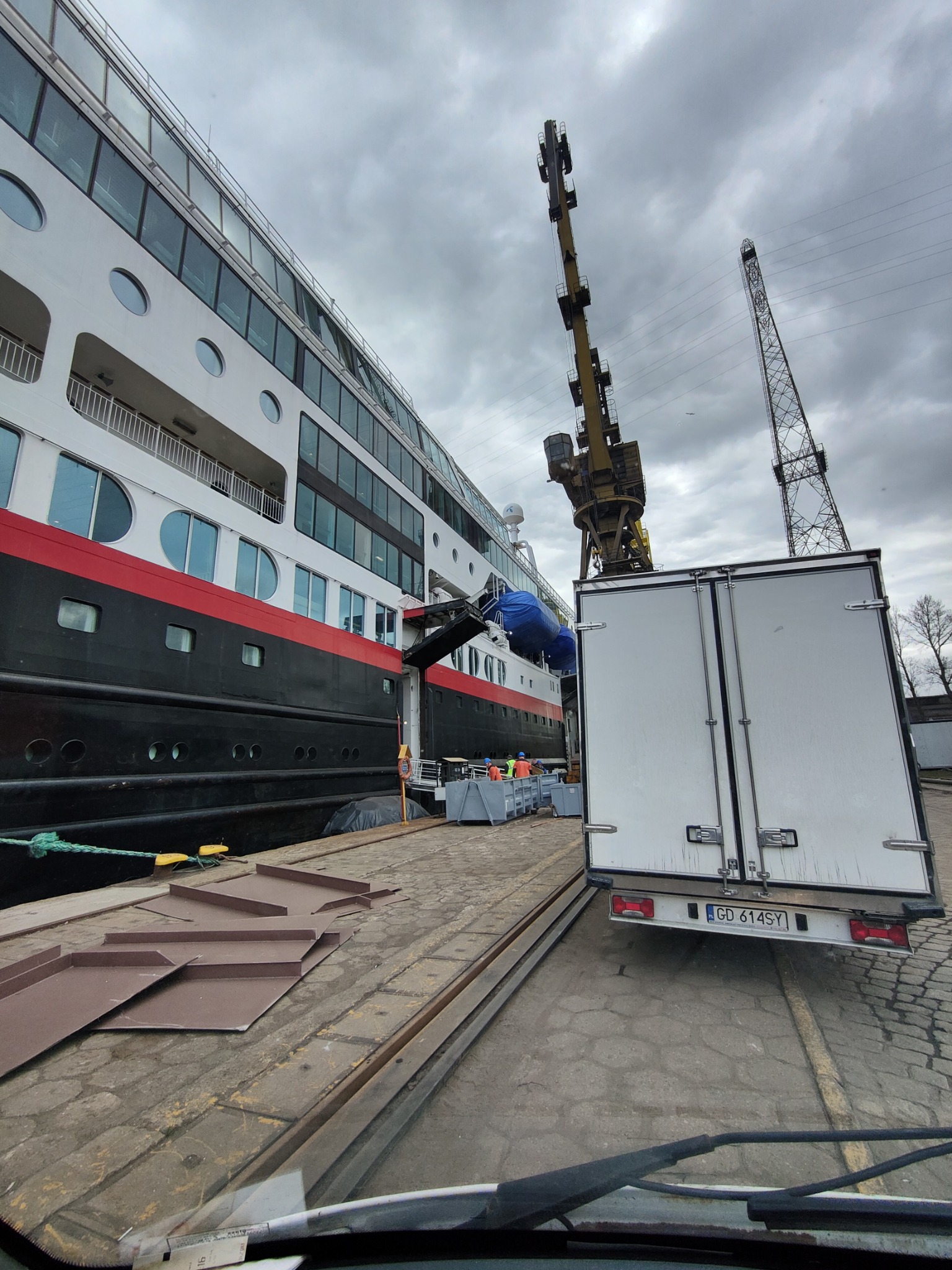 Ciężarówka dostawcza z otwartymi drzwiami zaparkowana obok statku wycieczkowego w porcie w Straszynie, z dźwigiem w tle i pracownikami w kamizelkach odblaskowych biorącymi udział w załadunku.