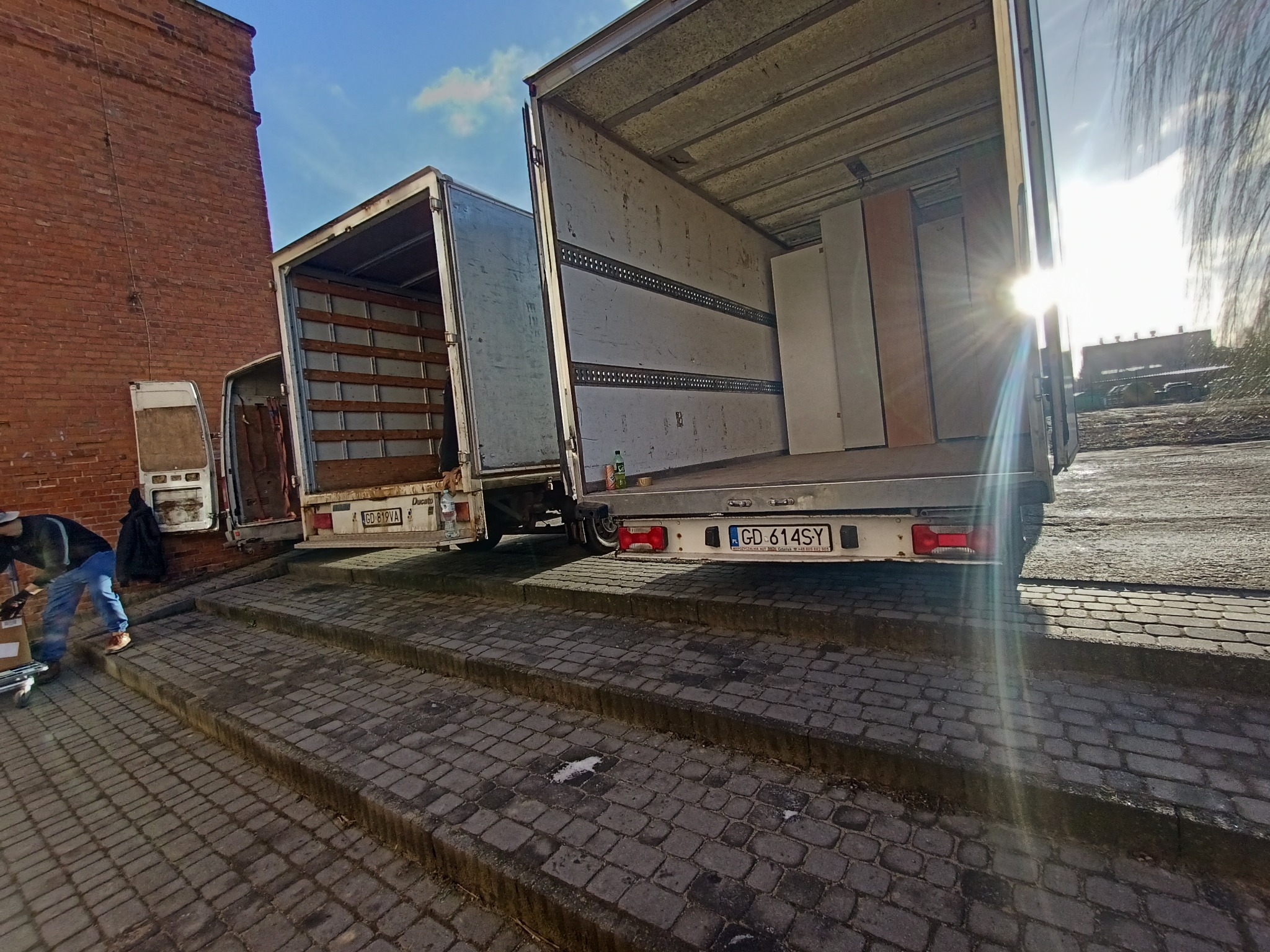 Trzy busy do przeprowadzek zaparkowane na schodach z kostki brukowej, jeden z pracownikiem w trakcie rozładunku, widok z dołu pod słońce.