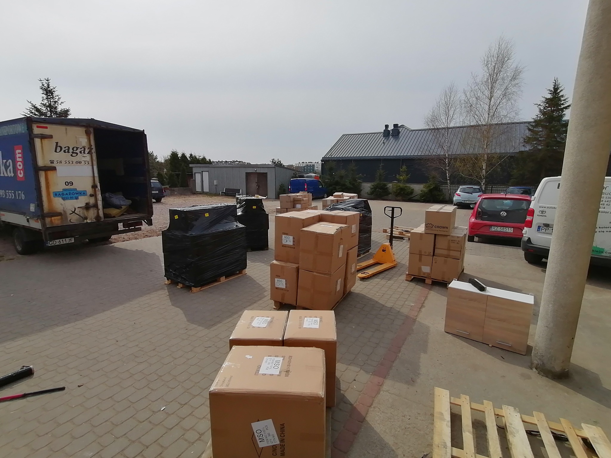 Częściowo załadowana ciężarówka z otwartymi drzwiami, obok palety z kartonami i meblem, na tle budynków gospodarczych w pochmurny dzień.