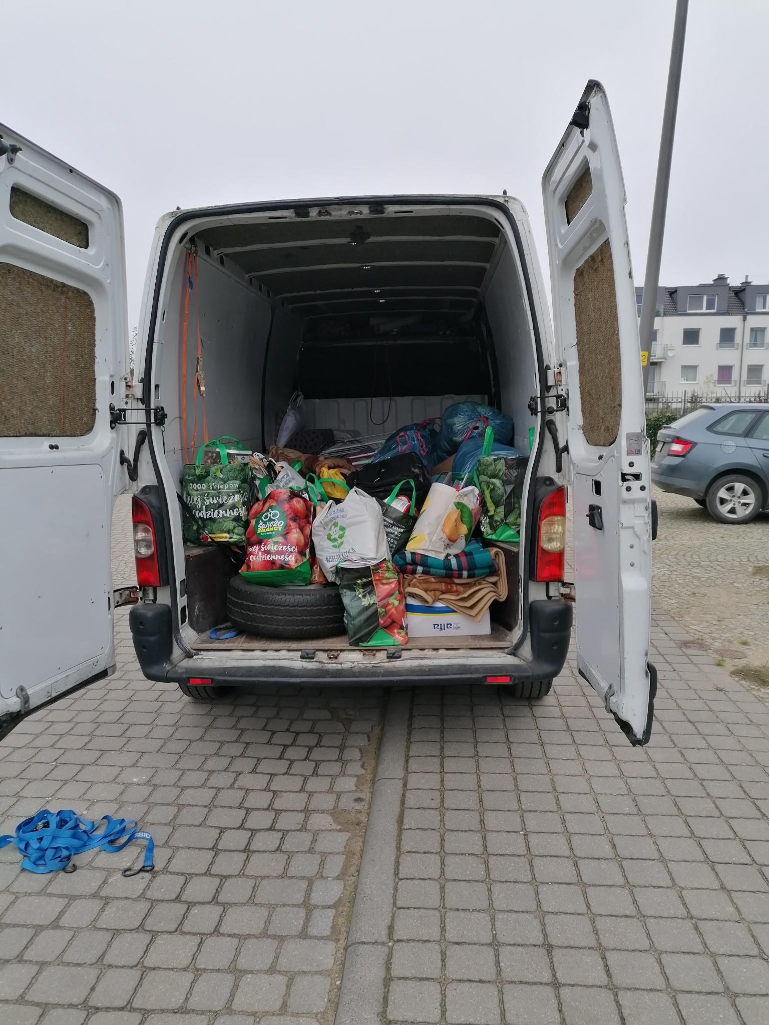 Wnętrze białego busa z otwartymi drzwiami załadowane torbami z zakupami, kocami i oponą, na zewnątrz kostka brukowa i niebieskie pasy transportowe.