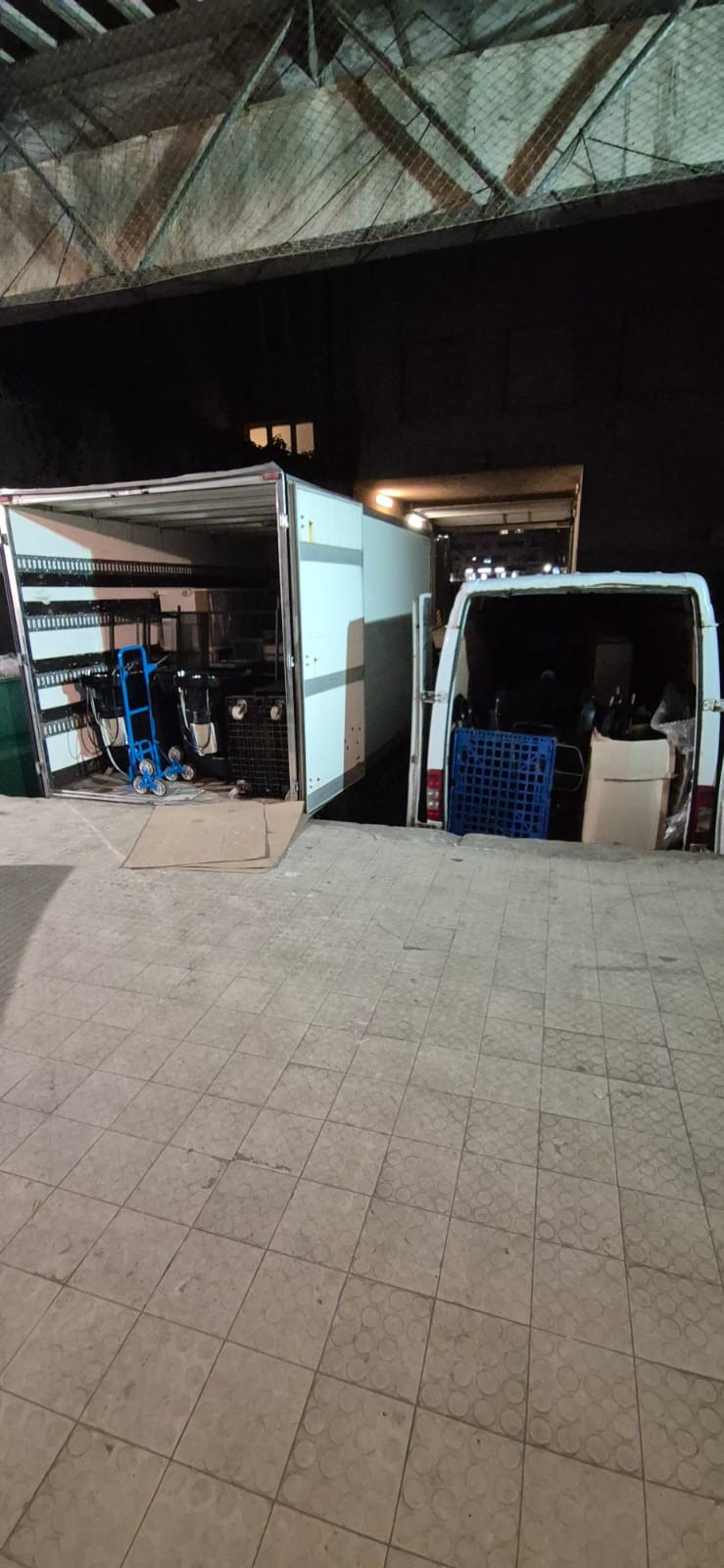 Dwa busy z otwartymi przestrzeniami ładunkowymi, jeden z wózkiem transportowym i sprzętem do przenoszenia szyb, drugi z niebieską skrzynką, na zewnątrz budynku w nocy.