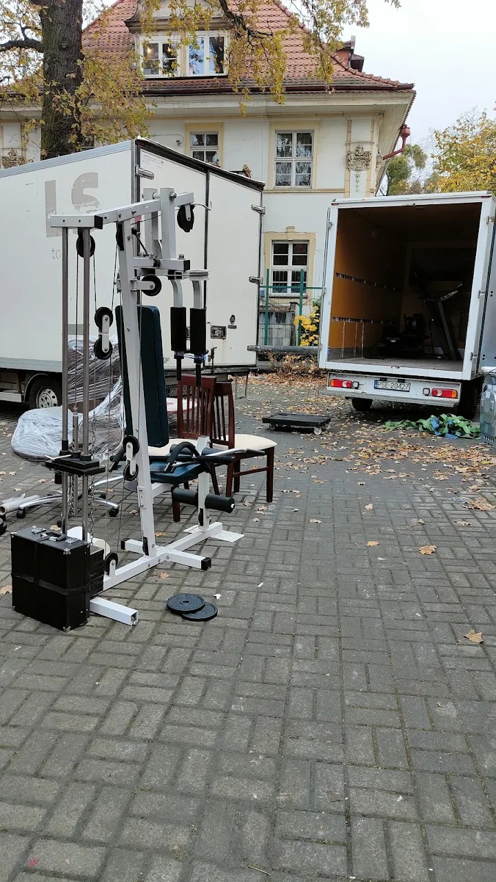 Sprzęt do ćwiczeń, krzesła i bieżnia wewnątrz otwartej furgonetki na tle budynku z czerwonym dachem i drzewa z opadającymi liśćmi.