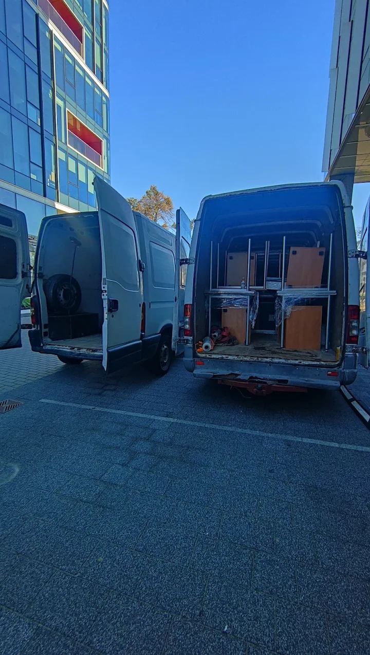 Dwa busy z otwartymi tylnymi drzwiami na parkingu; jeden z zapasowym kołem, drugi z meblami zabezpieczonymi folią wewnątrz przestrzeni ładunkowej.