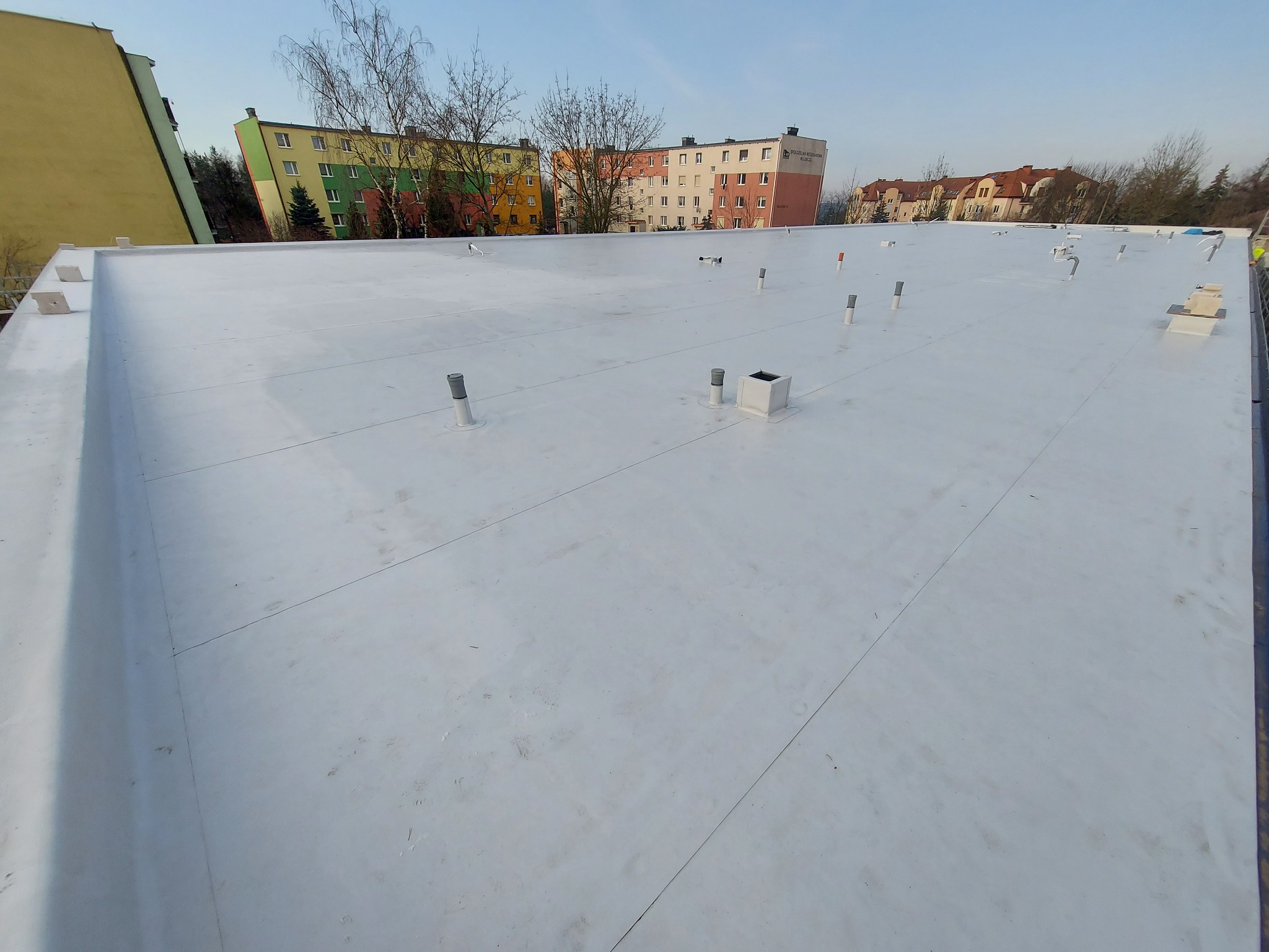 Biała, płaska powierzchnia dachu pokryta membraną hydroizolacyjną z widocznymi kominkami wentylacyjnymi i budynkami mieszkalnymi w tle.