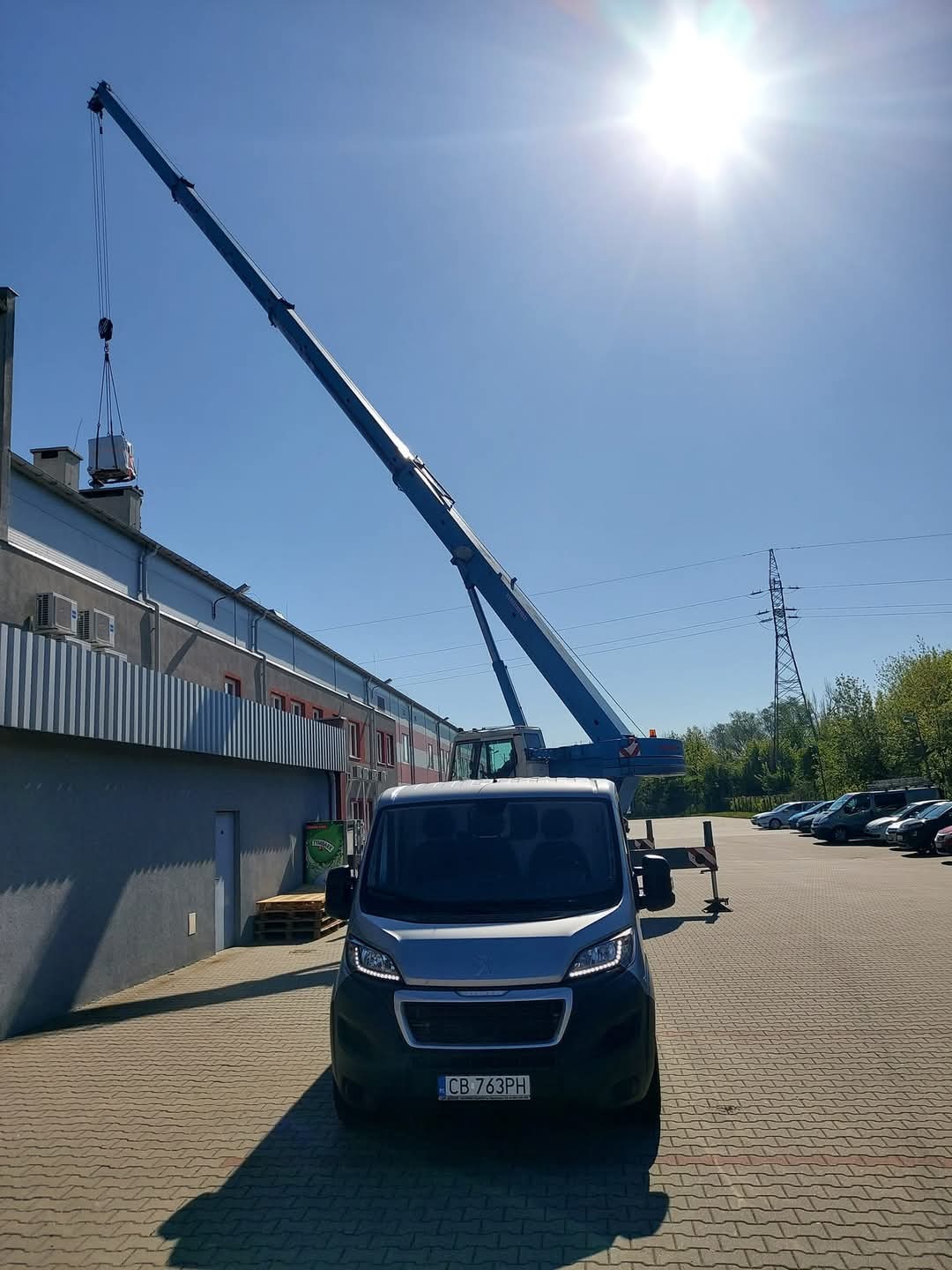Mobilny dźwig podnosi izolowany moduł na dach budynku przemysłowego, widoczny srebrny samochód dostawczy z przodu, błękitne niebo