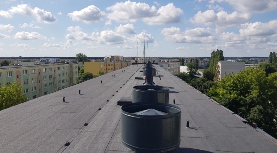 Widok z góry na płaski dach pokryty papą w Bydgoszczy, z widocznymi kominami wentylacyjnymi i budynkami mieszkalnymi w tle pod błękitnym niebem z chmurami.