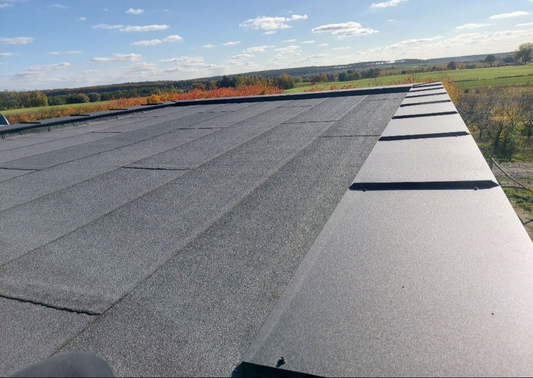 Dach pokryty szarą papą bitumiczną na tle jesiennego krajobrazu, widoczne obróbki blacharskie.