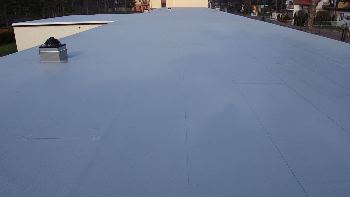 Rozległy, płaski dach pokryty szarą membraną dachową, z widocznym kominkiem wentylacyjnym i elementami wykończeniowymi, na tle zabudowy jednorodzinnej i drzew.