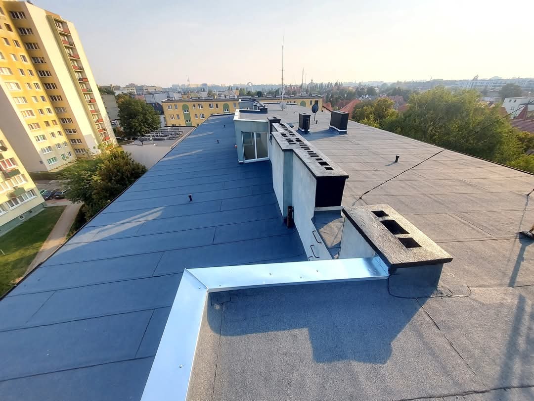 Rozległy, płaski dach pokryty papą termozgrzewalną w odcieniach szarości i czerni, z widocznymi elementami wentylacyjnymi i obróbkami blacharskimi z jasnego metalu, na tle zabudowy miejskiej...