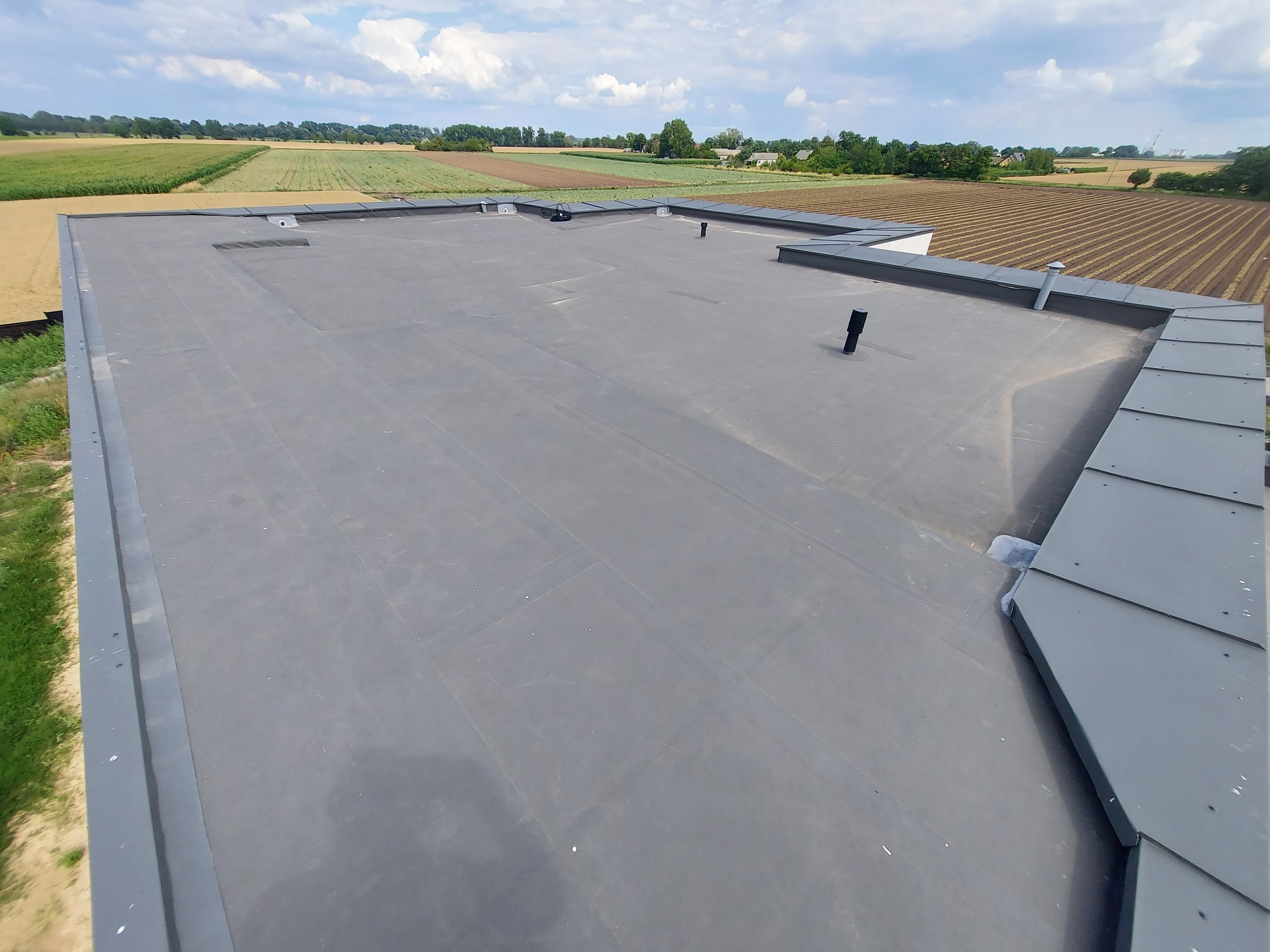 Szary dach płaski z widocznymi detalami obróbek blacharskich i kominkami wentylacyjnymi. W tle pola uprawne pod błękitnym niebem.