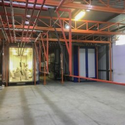 TECHNIKOR SPÓŁKA Z OGRANICZONĄ ODPOWIEDZIALNOŚCIĄ - Wnętrze hali produkcyjnej z kabiną lakierniczą, gdzie dwóch pracowników w kombinezonach ochronnych przygotowuje metalowe elementy do nałożenia farby. Widoczne zawieszone detale i instalacja...