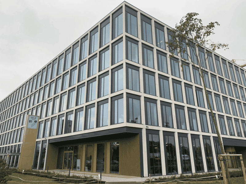 Nowoczesny budynek biurowy z prostokątną fasadą, licznymi oknami w ciemnych ramach i elementami elewacji w kolorze jasnego drewna, widoczny znak informacyjny przy wejściu.