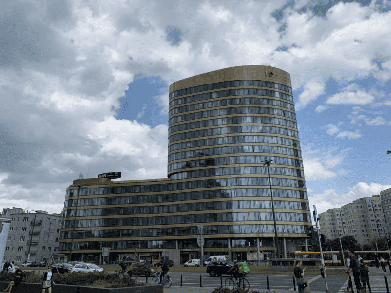 Wysoki budynek biurowy z zaokrąglonymi narożnikami, wykończony w odcieniach złota, widoczny z perspektywy ulicy w słoneczny dzień z chmurami w tle.