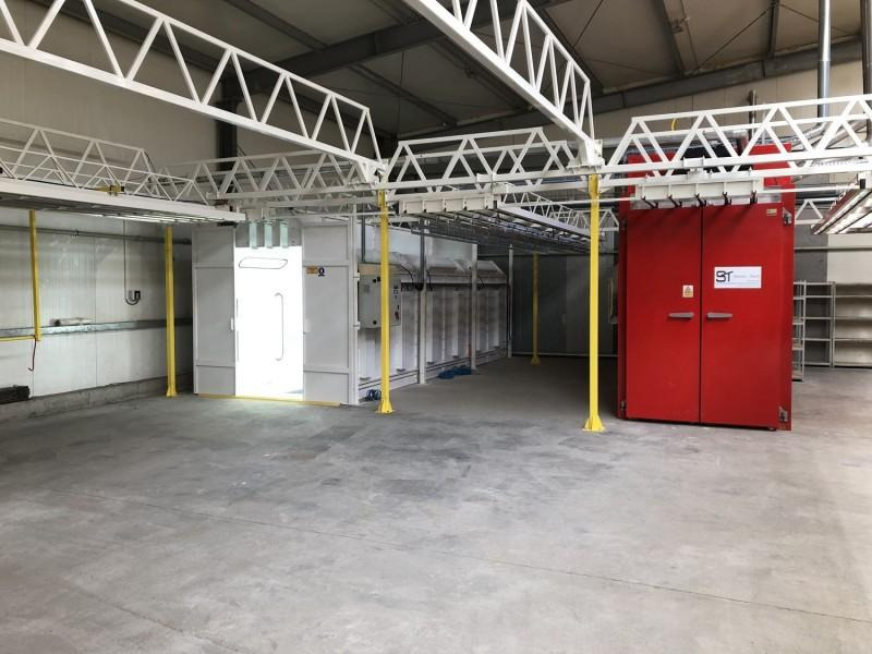 Wnętrze hali z linią technologiczną do obróbki powierzchniowej metali, widoczna kabina lakiernicza z otwartymi drzwiami i piec w kolorze czerwonym.