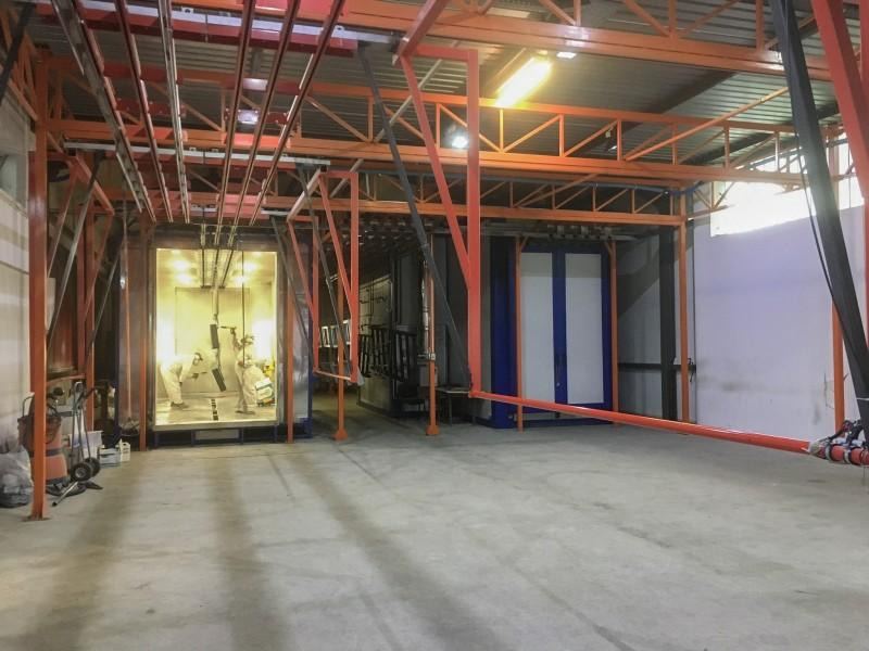 Wnętrze hali produkcyjnej z kabiną lakierniczą, gdzie dwóch pracowników w kombinezonach ochronnych przygotowuje metalowe elementy do nałożenia farby. Widoczne zawieszone detale i instalacja...