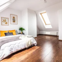 Jasne poddasze z drewnianą podłogą i oknem dachowym. Ustawione łóżko z roślinami po bokach. Minimalistyczne plakaty na ścianie dodają uroku.
