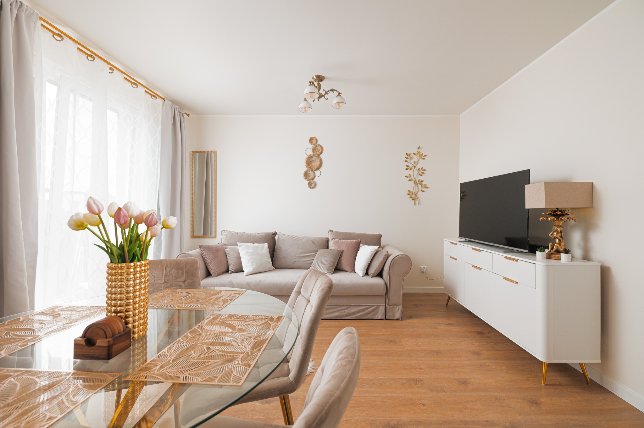 Jasne wnętrze salonu z kanapą, szklanym stołem z kwiatami i białym kredensem z TV. Neutralna kolorystyka, złote akcenty dekoracyjne. Przytulna aranżacja.